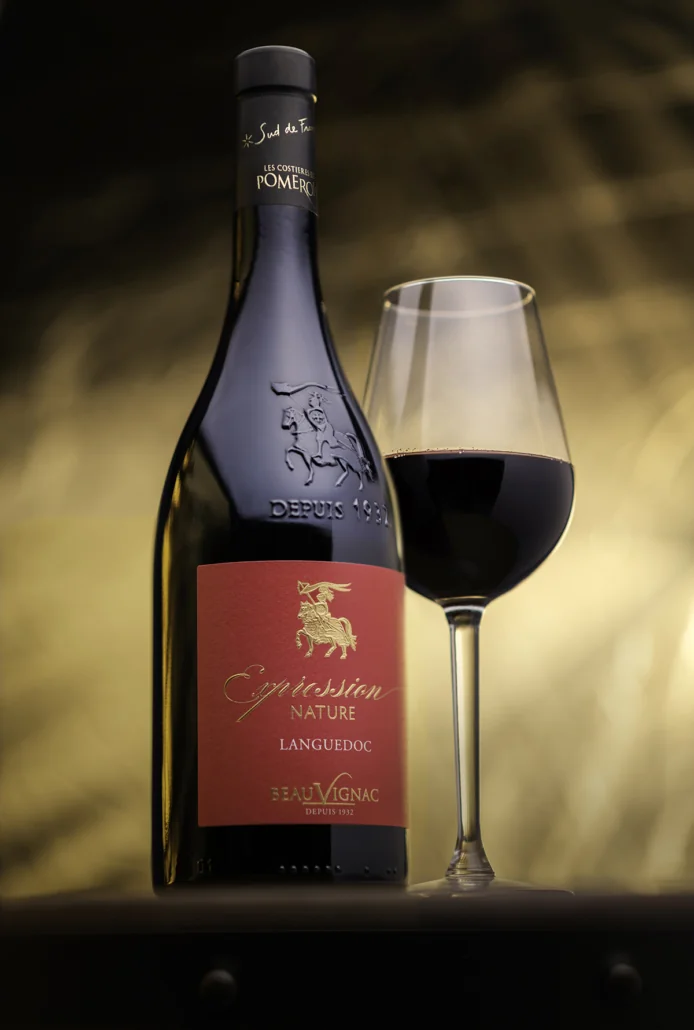 Packshot lifestyle haut de gamme : bouteille 'Expression Nature Languedoc' (etiquette rouge, blason chevalier en relief sur le verre) avec un verre de vin rouge, fond chaud dore en bokeh. Composition soignee, lumiere chaleureuse.