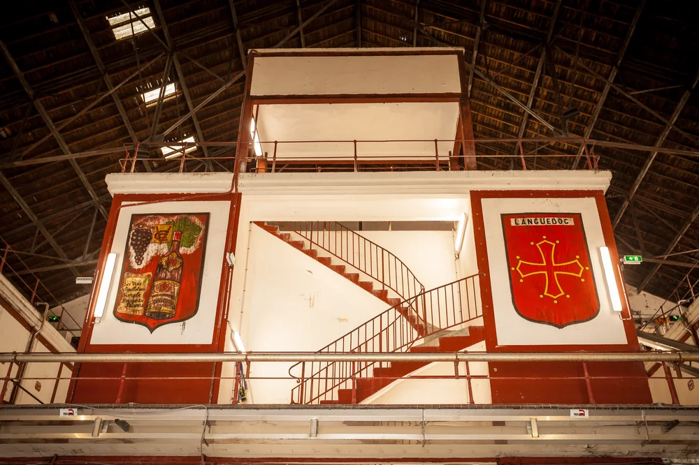 Intérieur de la halle historique de la coopérative : charpente bois en voûte, escalier tournant et deux fresques peintes (bouteille de vin et croix occitane du Languedoc). Architecture patrimoniale forte.