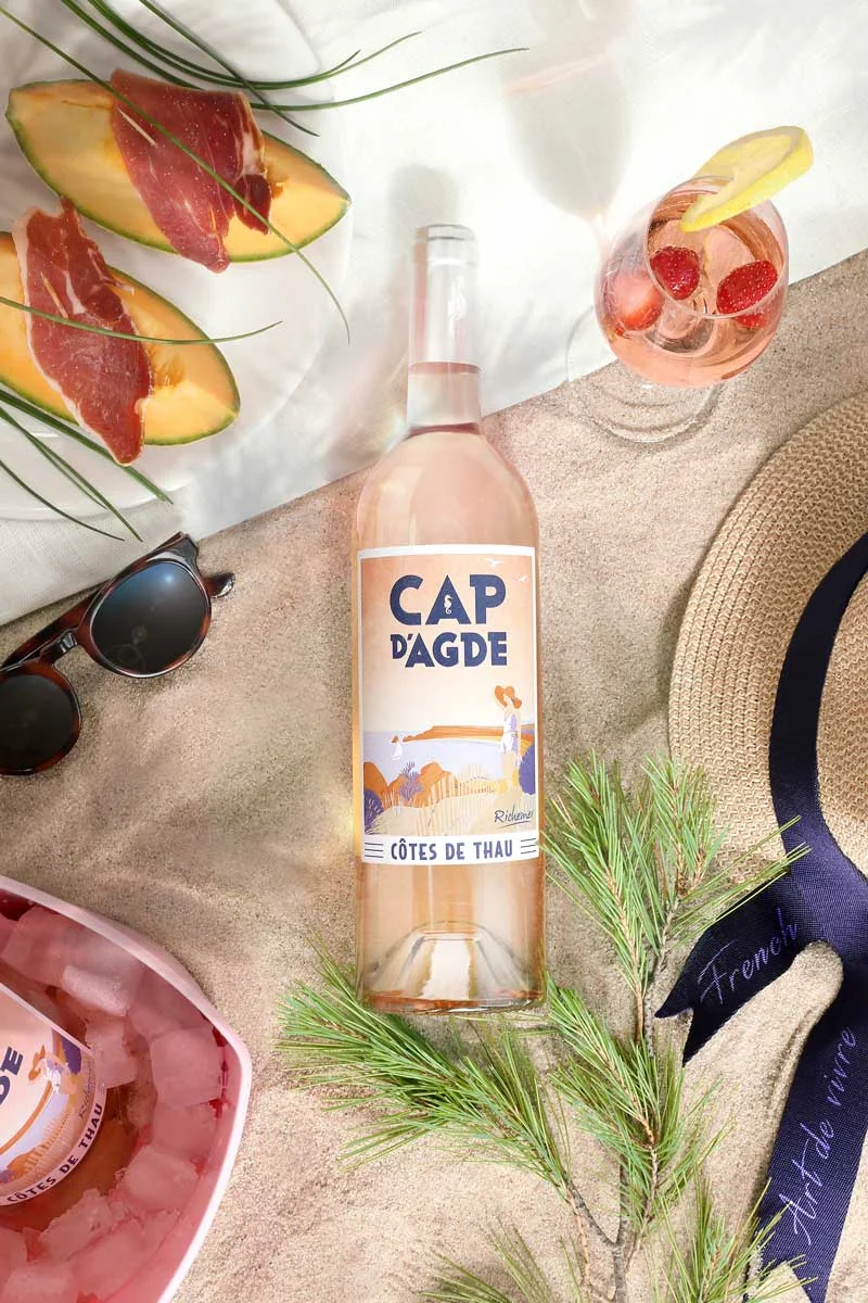 Mise en scene estivale art de vivre : bouteille de rose Cap d'Agde posee sur le sable avec lunettes, chapeau de paille, melon-jambon et verre de rose. Lifestyle premium, tres vendeur.