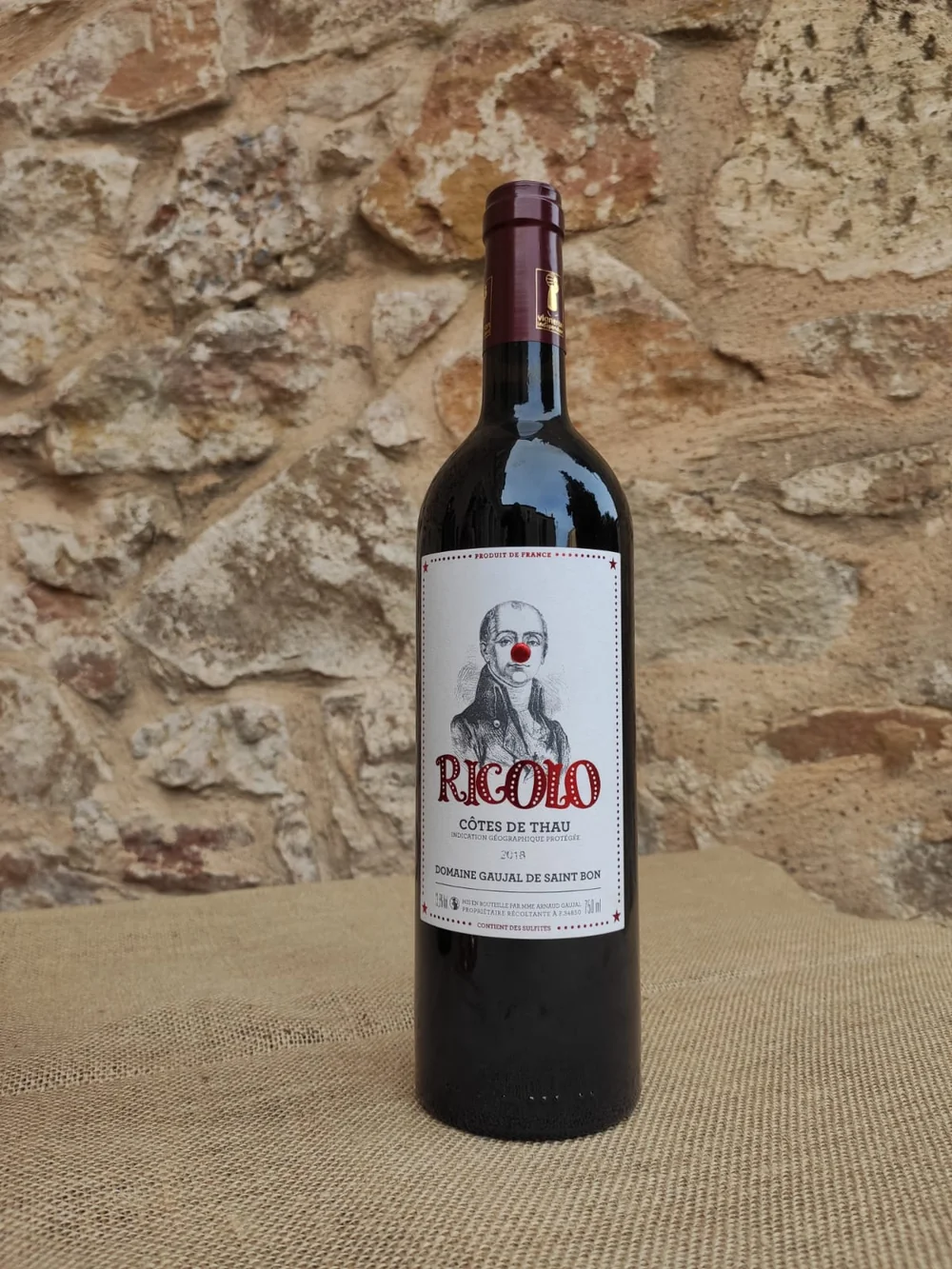 Packshot lifestyle : bouteille de rouge 'RIGOLO' Cotes de Thau (Domaine Gaujal de Saint Bon) posee sur toile de jute devant un mur de pierres seches. Belle mise en situation. 1200x1600.