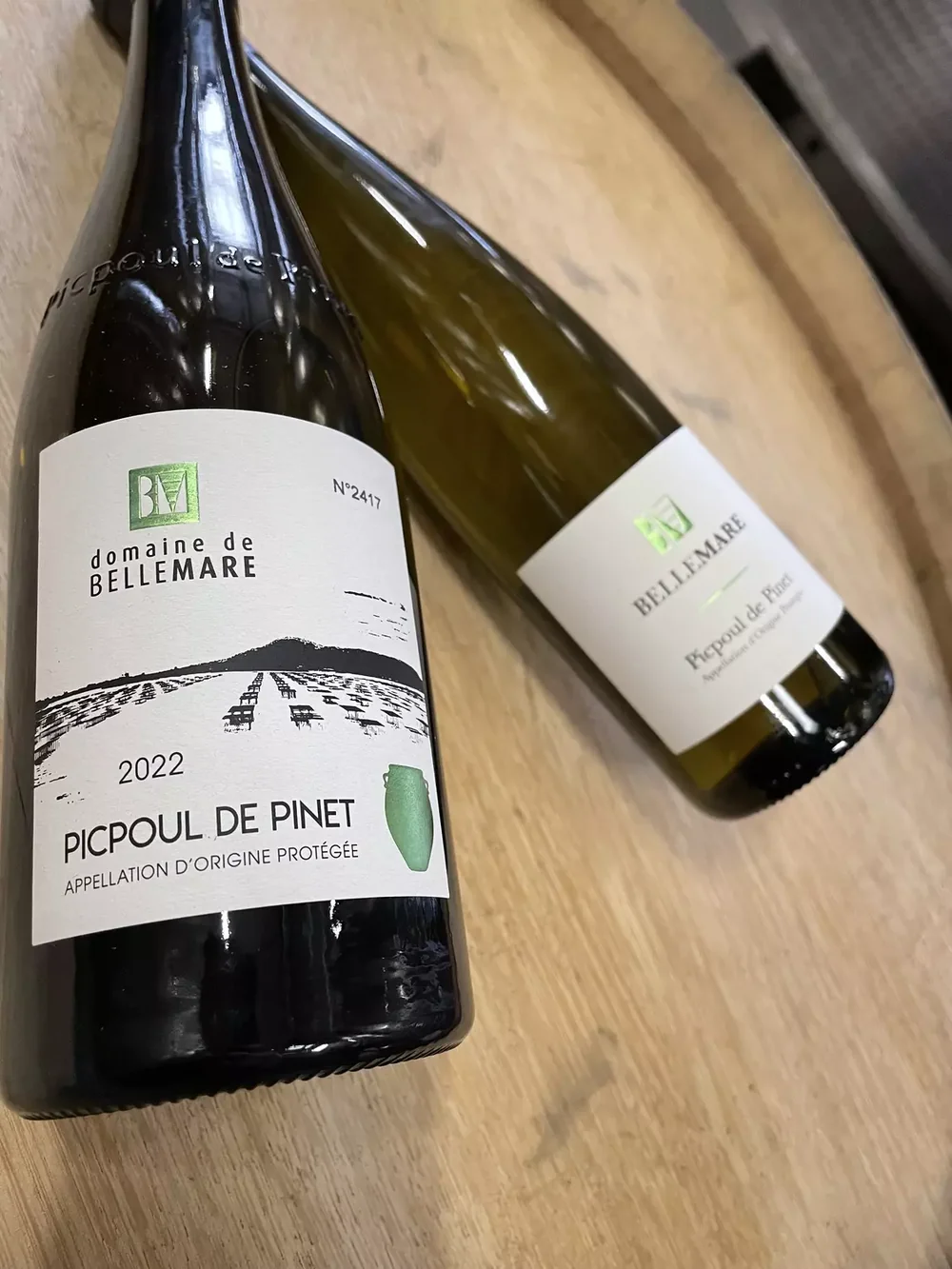 Deux bouteilles de Picpoul de Pinet du domaine posées sur un tonneau, étiquette bien lisible, lumière naturelle. Belle mise en situation, pas un packshot plat.
