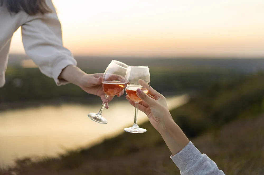 Scene lifestyle de degustation : deux mains trinquent avec des verres de rose au coucher du soleil au-dessus d'un plan d'eau / paysage flou, lumiere doree. Image conviviale et evocatrice, tres valorisante pour suggerer l'art de vivre autour du vin.