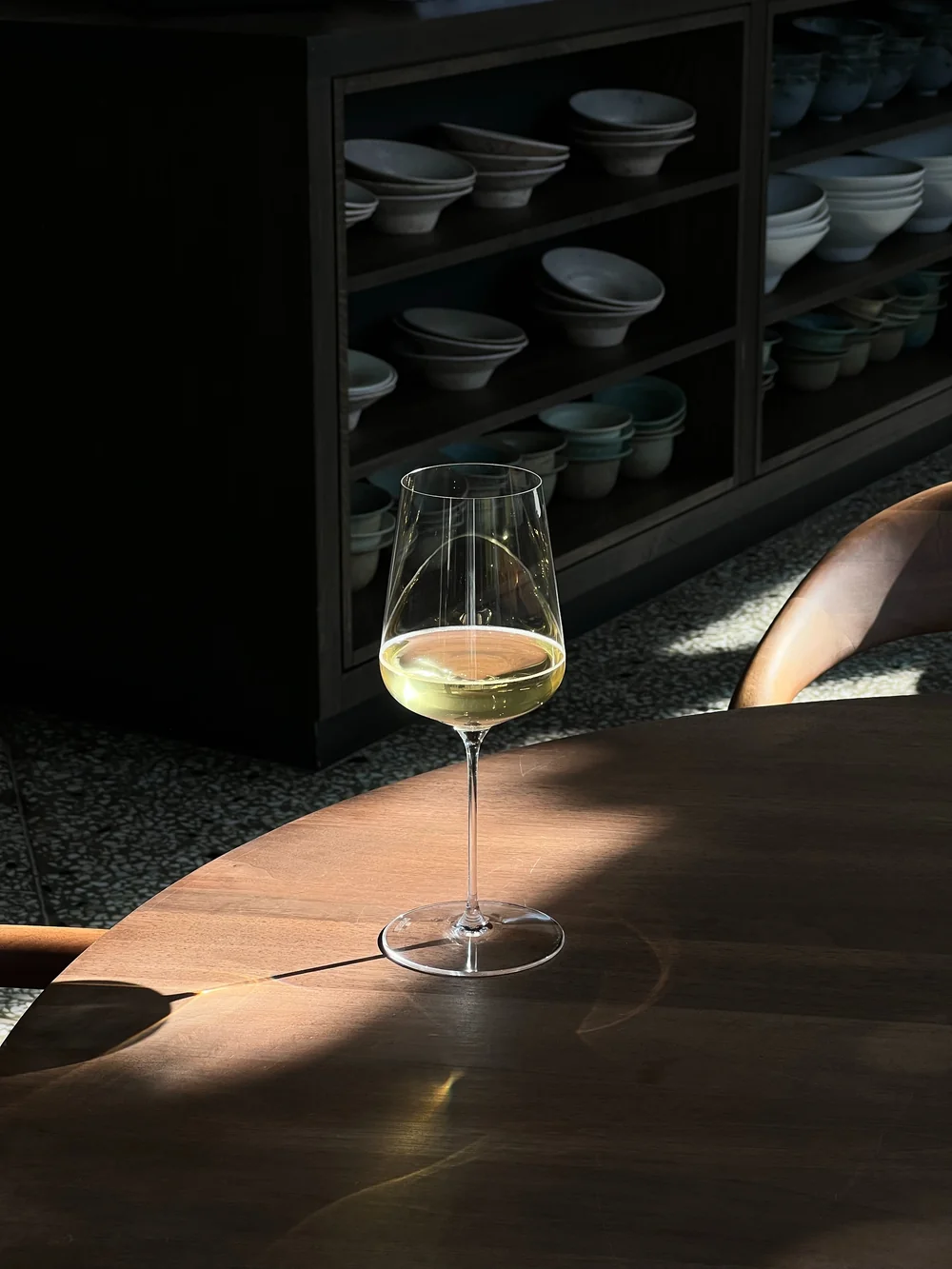 Photo lifestyle elegante : un verre de vin blanc pose sur une table ronde en bois sombre, baigne d'un rai de lumiere, avec en arriere-plan un meuble noir garni de bols en ceramique. Composition raffinee, ambiance contemporaine et premium (mise en situation, pas une etiquette plate).